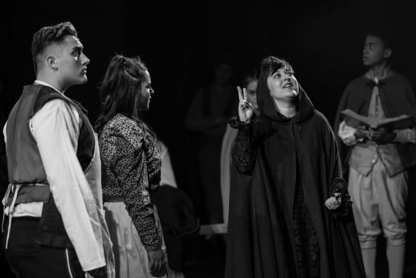 cast gathered around the witch who is looking up into the sky
    
