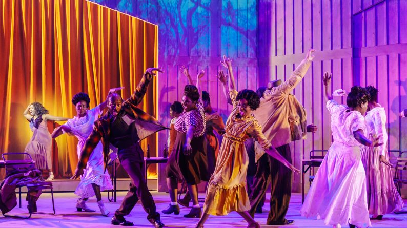 Production image from The Color Purple. The company dance in pairs on a purple and blue toned stage, with a golden curtain in the background.