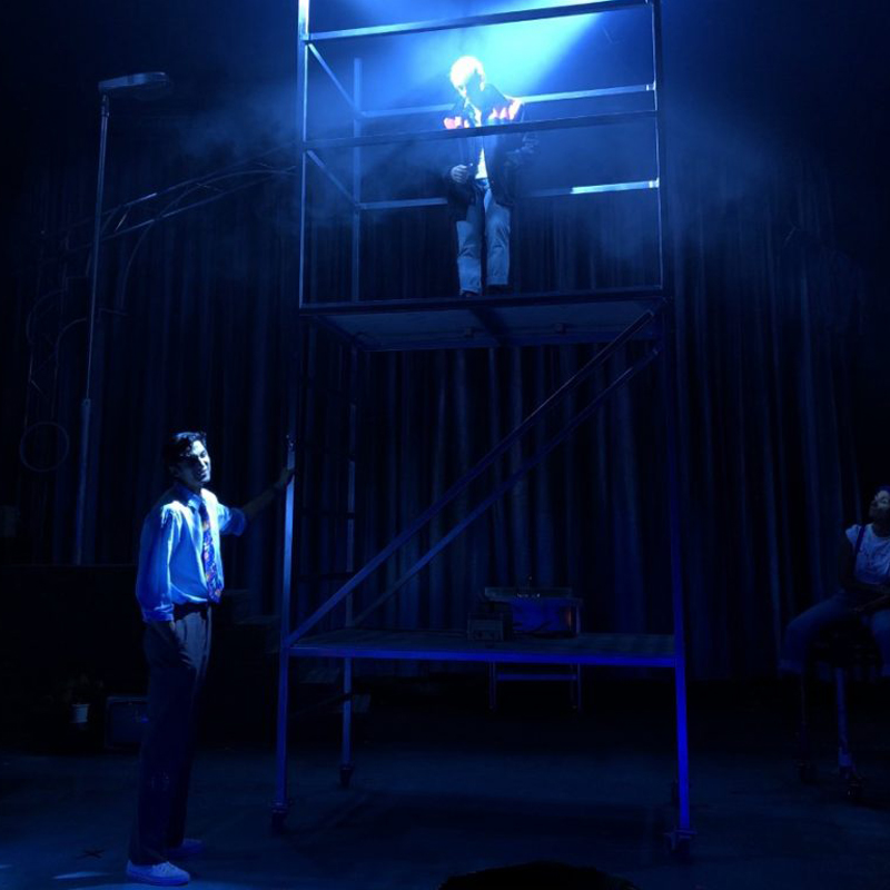 Jonny Fines as Johnny looks down from a platform at Omar Malik as Omar in a candid image from My Beautiful Laundrette rehearsals. The stage is illuminated in a wash of blue light and haze.