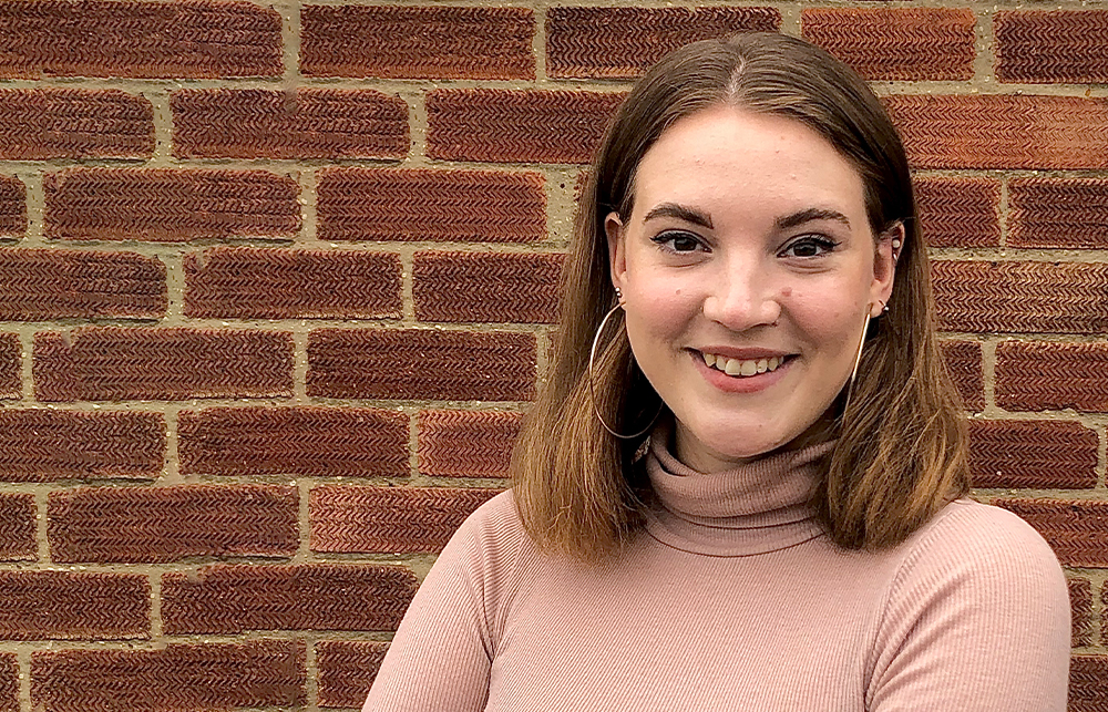 Jennifer Lane Baker smiles in front of a brick wall. She is wearing a dusty pink polo neck, with hoop earrings and her hair down.