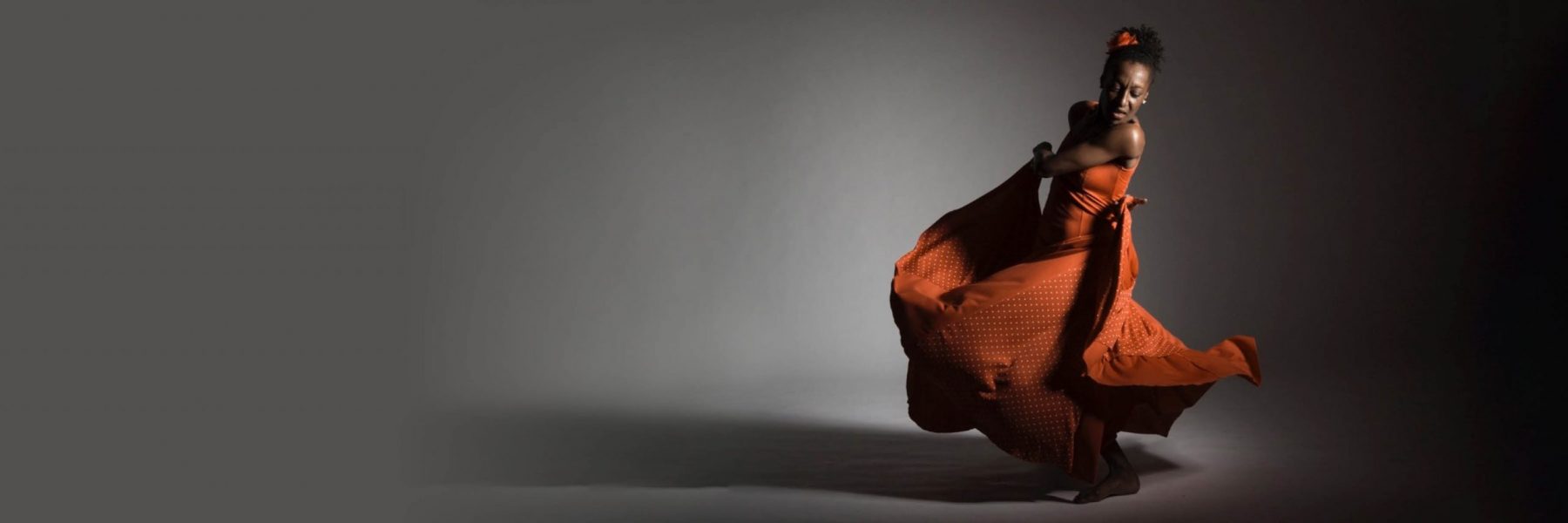 Dollie Henry dances in front of a grey backdrop. She is turned slightly away from the camera, looking down, as she swishes an orange dress.
    