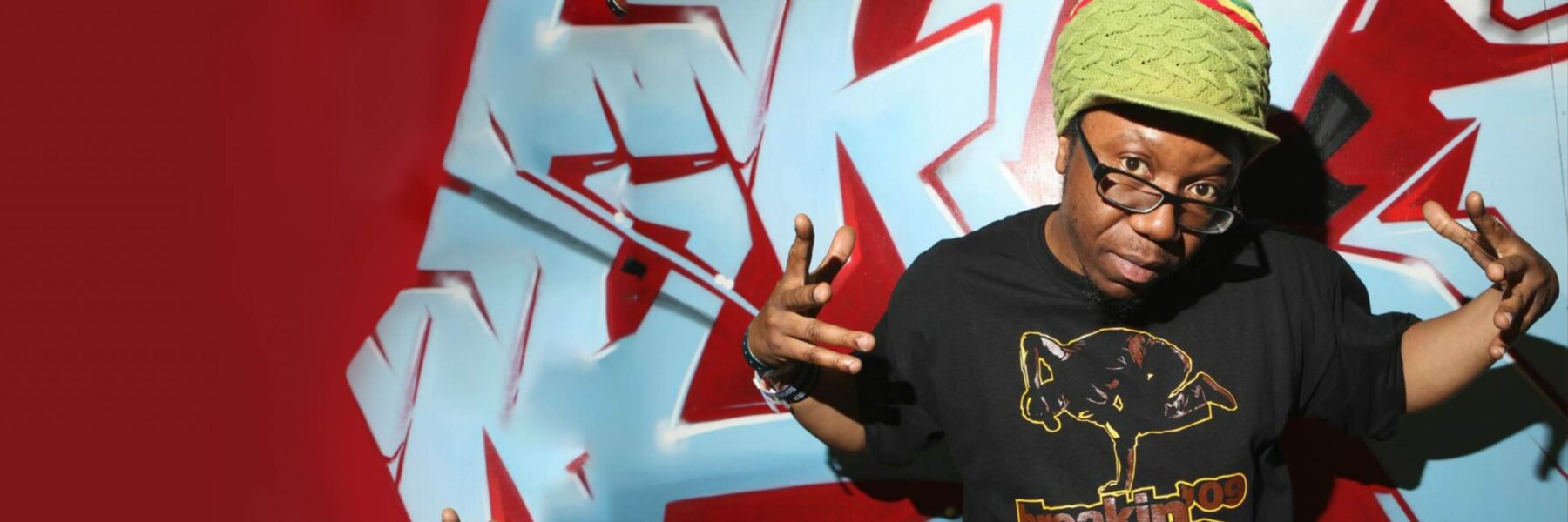 Jonzi D smiles with his hands pointing towards the camera. Pictured from the waist up in front of a graffiti wall, he is wearing a slogan top and green hat, with black glasses.
    