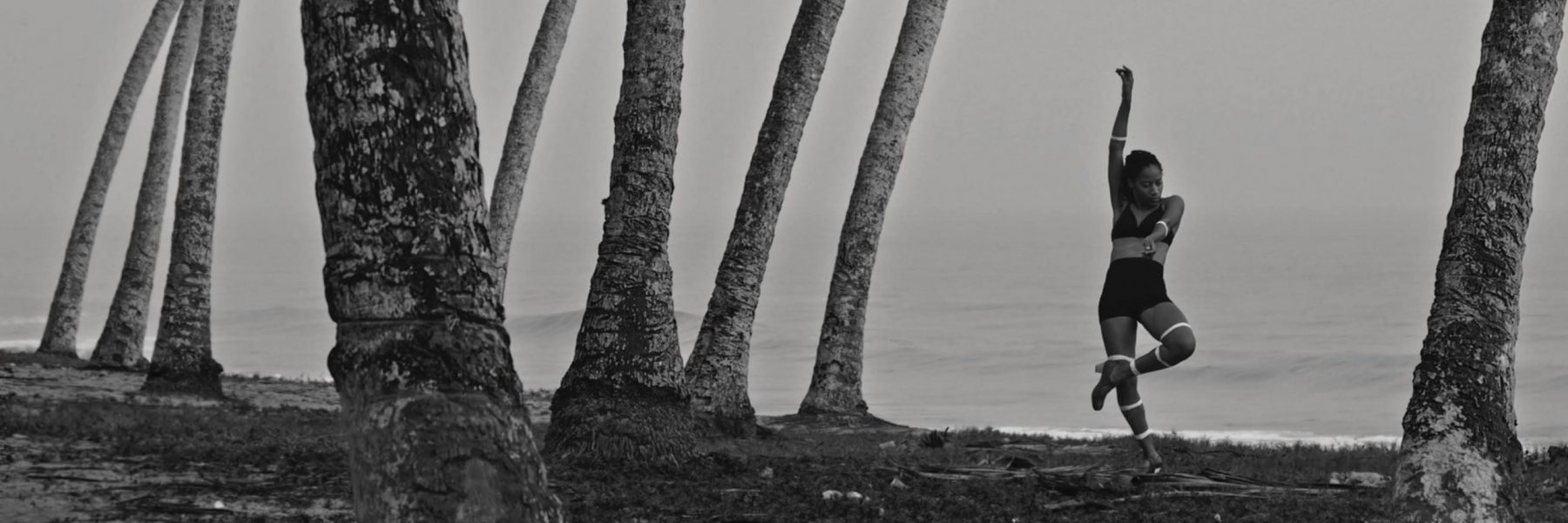 A black and white image of Yinka Esi Graves dancing beneath trees on a shoreline. She is wearing a black bralette and skirts with light-coloured heels. She is standing on her right foot with her right arm in the air, her left arm and leg crossing her body.
    