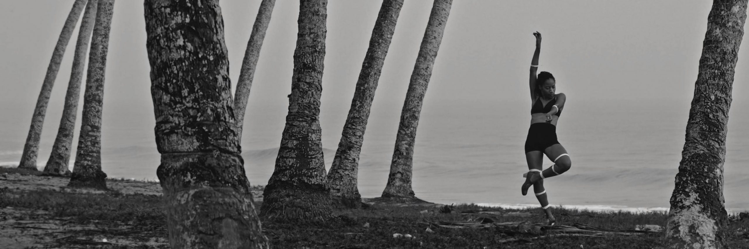 A black and white image of Yinka Esi Graves dancing beneath trees on a shoreline. She is wearing a black bralette and skirts with light-coloured heels. She is standing on her right foot with her right arm in the air, her left arm and leg crossing her body.