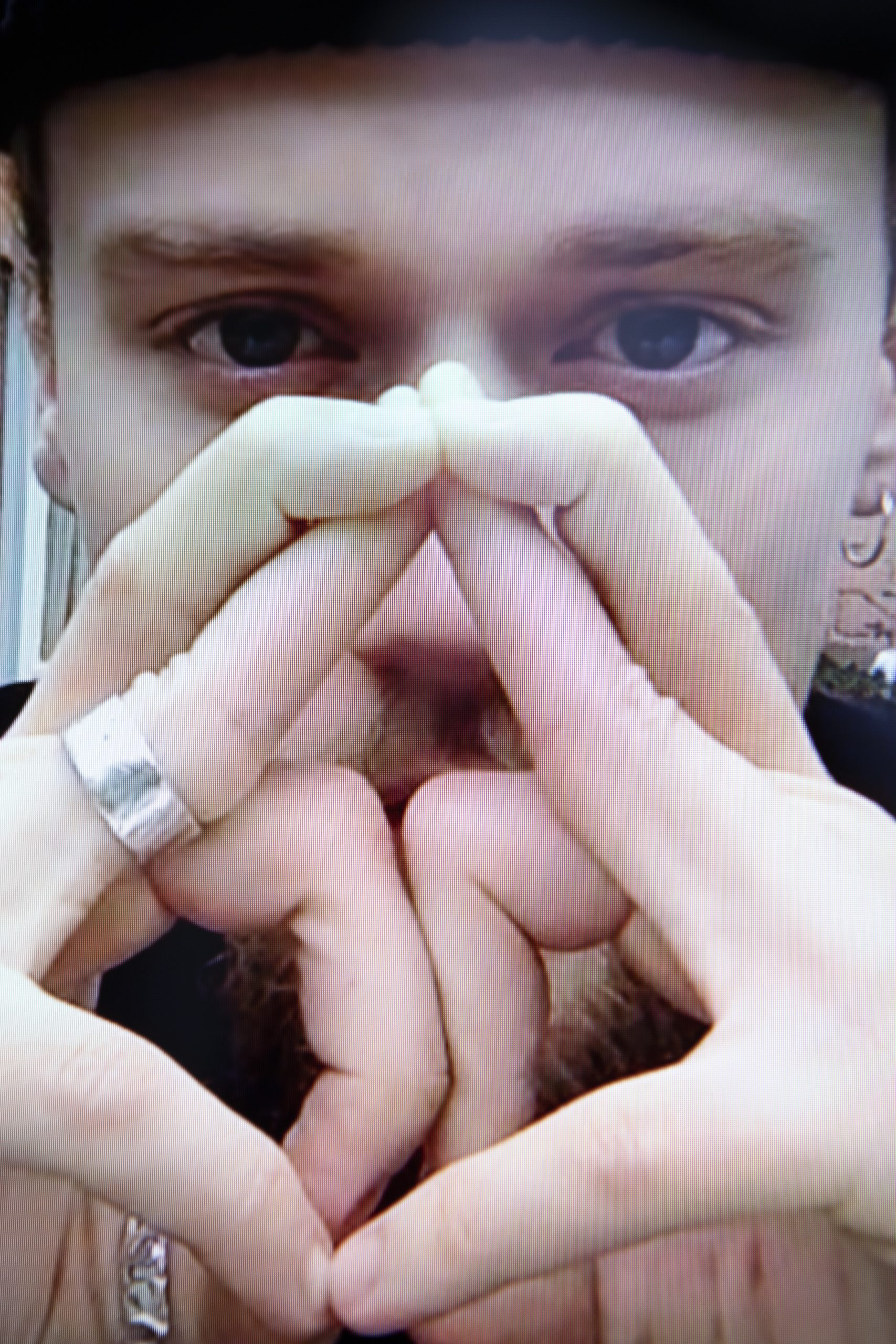 Hal Mayer is a white man with dark blue eyes. He stares into the camera in this close-up image with his hands in front of his face, fingers entwined into triangular shapes. He wears a wide silver ring on a finger on his right hand.