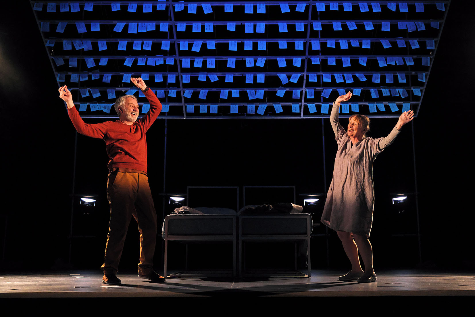 Production image from Maggie May. Maggie (Eithne Brown) and Gordon (Tony Timberlake) dance with glee around their bed beneath a ceiling covered in blue post-it notes, their top halves illuminated in warm spotlights.