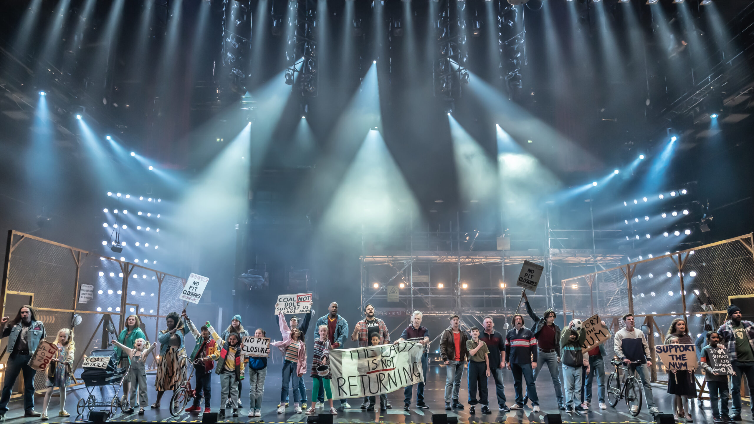 Production image from Billy Elliot The Musical. The community are gathered on the picket line for the Miners' Strike, in dark, moody lighting.