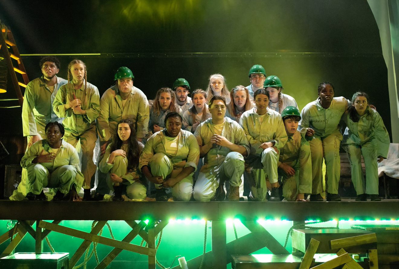 Production image from Oh What A Lovely War. The company huddle together in light blue boiler suits and green hard hats. They look off into the distance, illuminated in green light,