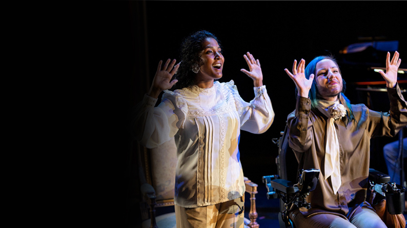 An Asian woman with long dark stands, with her hands held up, next to a person using a wheelchair, also with their hands held up, with dark and blue hair. Both smile.