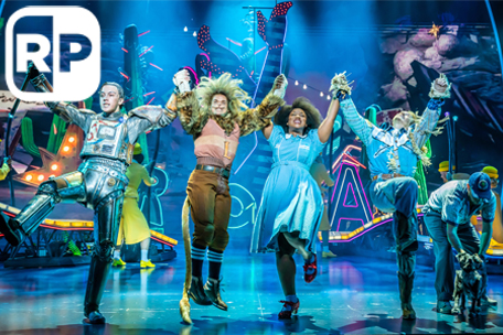 Production photograph from The Wizard of Oz. Tin Man (Paul French), Lion (Giovanni Spanó), Dorothy (Georgina Onuorah), Scarecrow (Jonny Fines) and Toto (Ben Thompson) leap into the air joyfully in front of neon signs on a blue stage. There is a white RP - Relaxed Performance - logo top left).