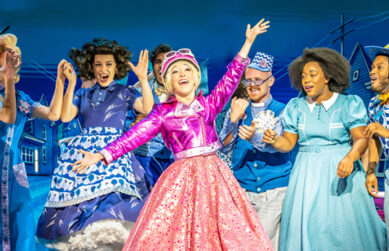 Production image from The Wizard of Oz. Glinda the Good Witch - wearing a shiny pink jacket, a sparkly pink a-line skirt and a pink helmet with a blonde ponytail - exclaims with her arms aloft. Four munchkins and Dorothy, all wearing blue toned outfits, watch on with joyful expressions.