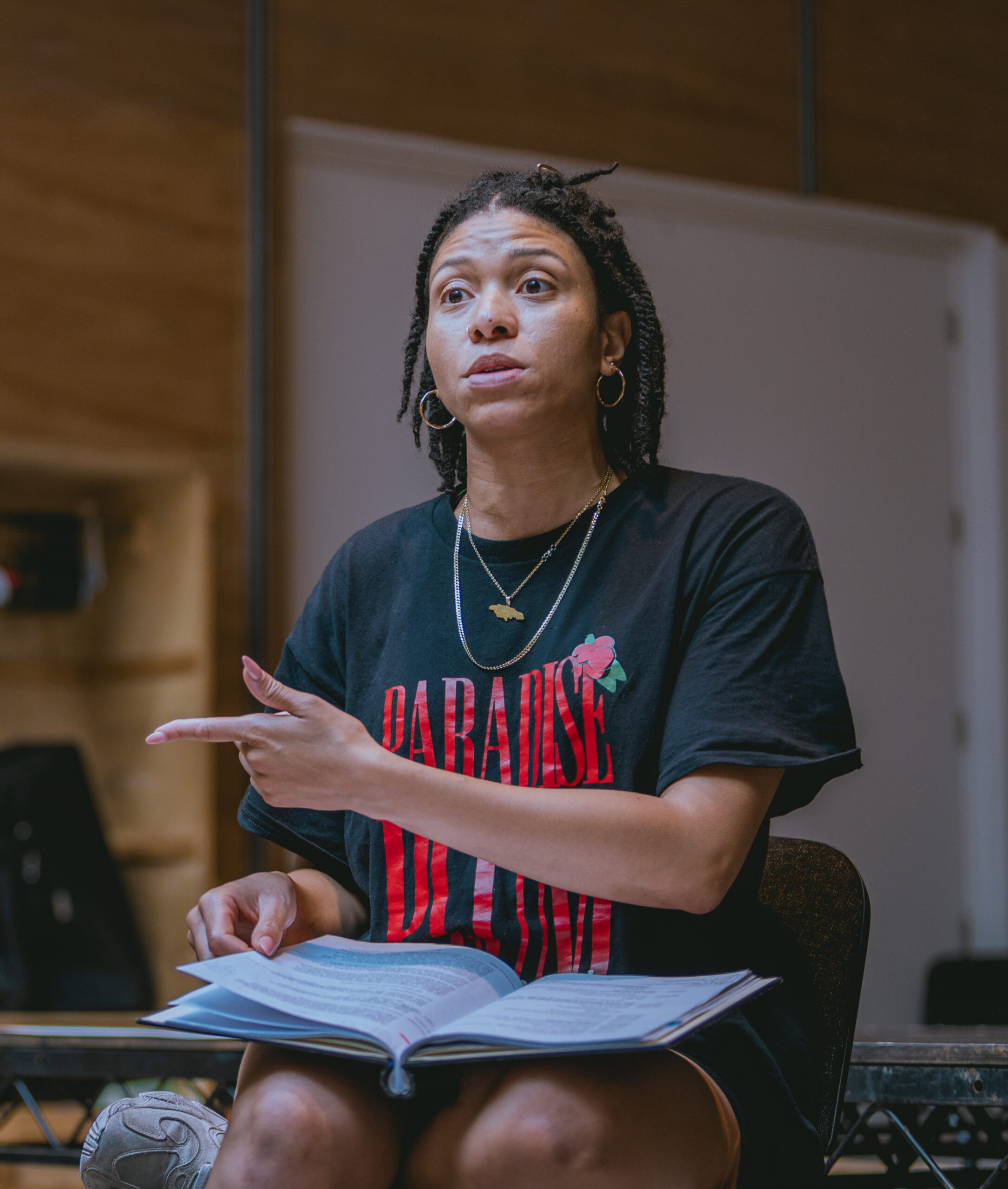 In the rehearsal room, Hannah Sinclair Robinson (Grete) is speaking to someone in the distance, pointing to the right with her left hand and with her script in her lap. She is wearing a black top with a red slogan.