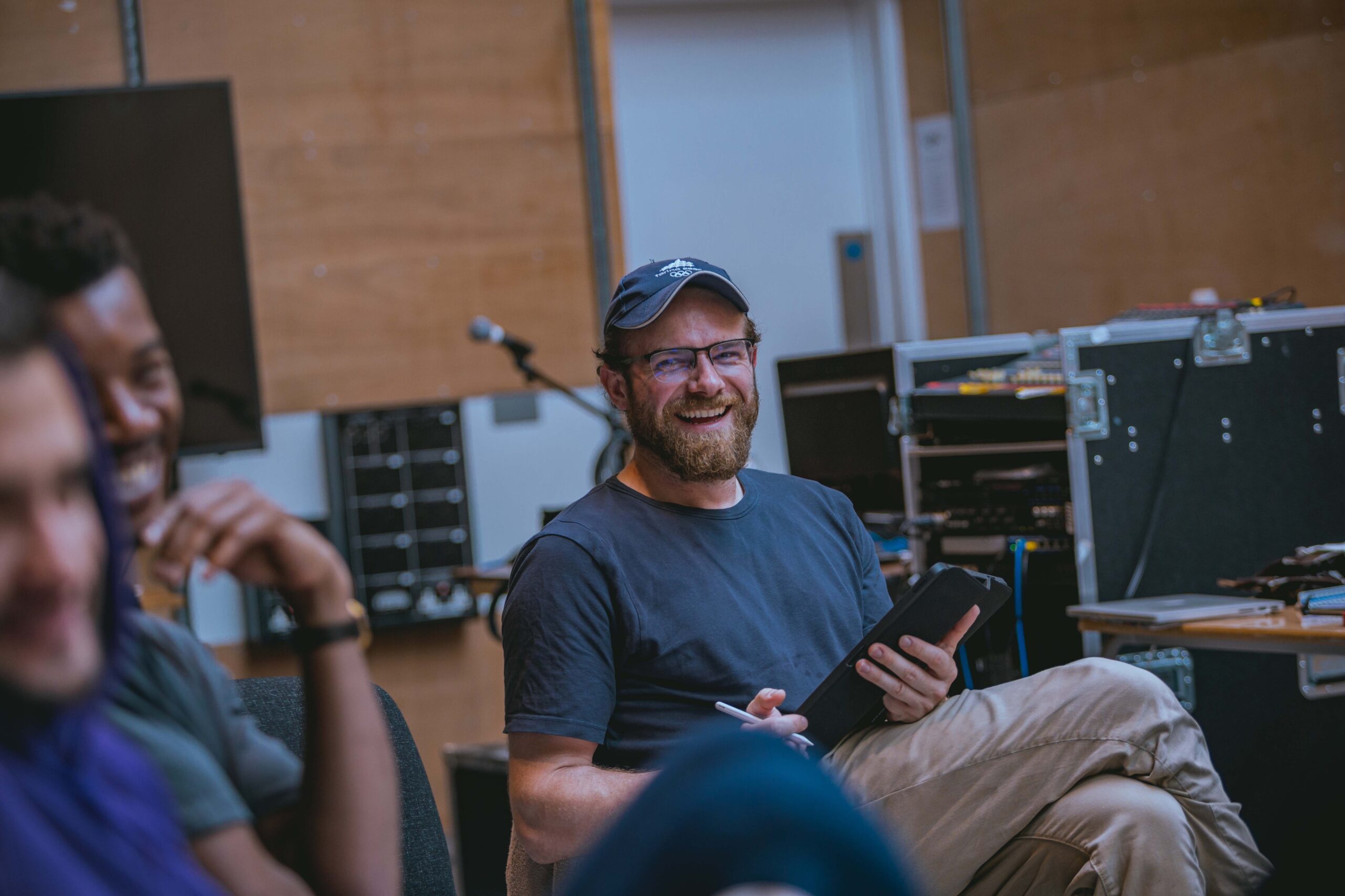 In a wood-cladded rehearsal room, Joe Layton (Chief Clerk/Lodger) is smiling as his sits with an electronic tablet in his left hand and smart pencil in his right. Blurred in the foreground are cast members Felipe Pacheco (Gregor) and Troy Glasgow (Father) who are also smiling. Several flight cases can be seen behind them.