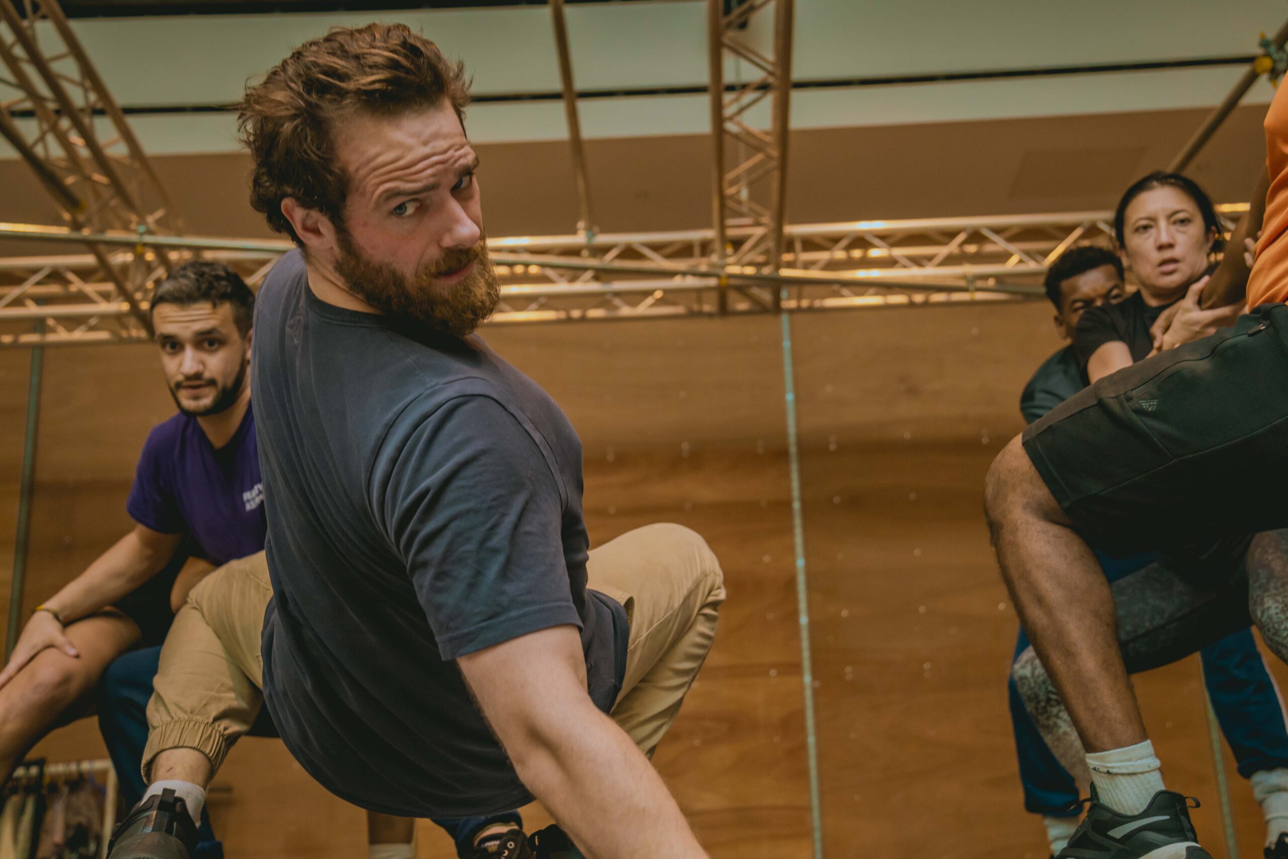 In a wood-cladded rehearsal room, the cast of Metamorphosis are rehearsing a variety of positions. In the foreground is Joe Layton (Chief Clerk/Lodger) who is looking over his right shoulder, almost falling into the camera as the other cast members watch on. All are wearing casual leisurewear.