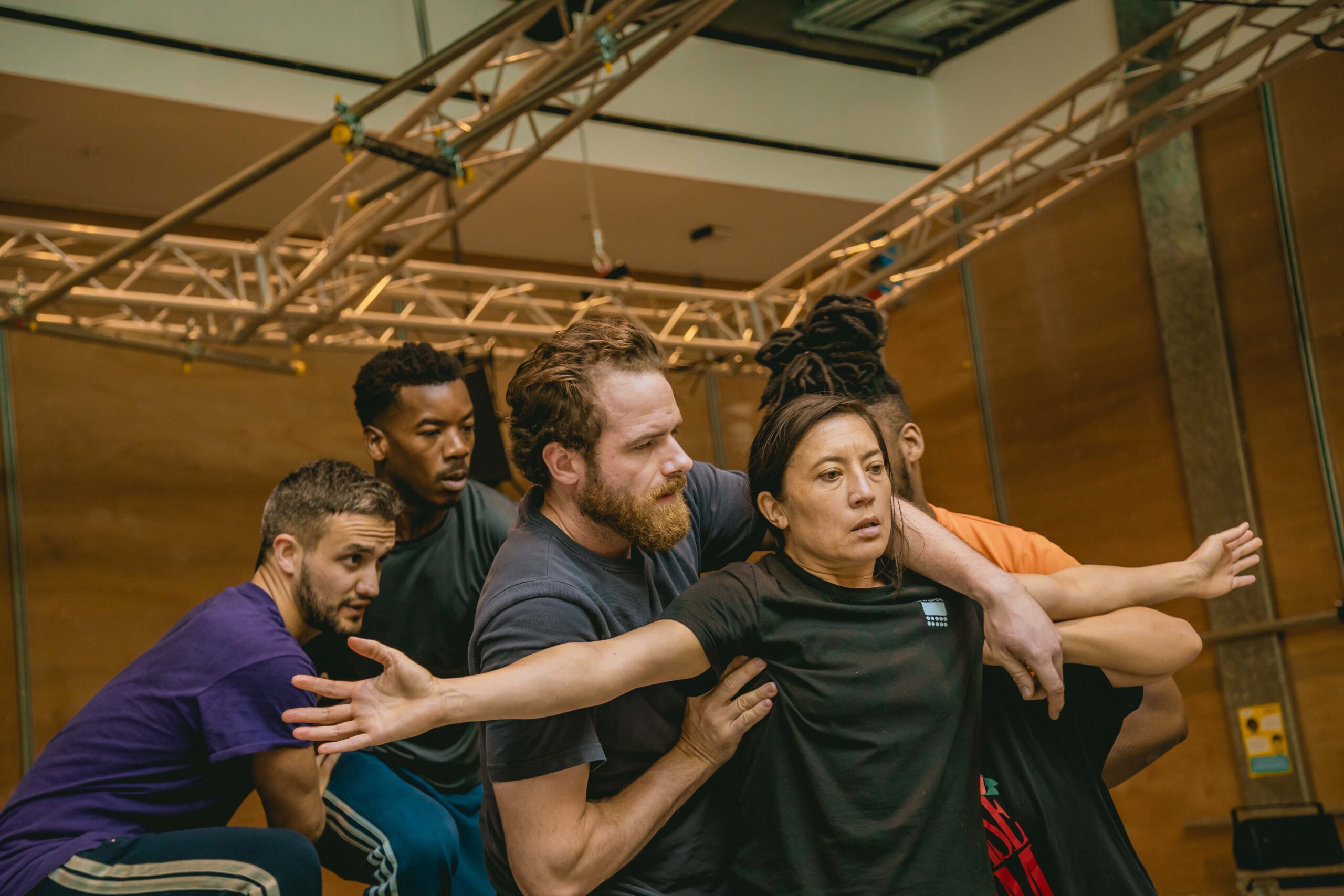 In a wood-cladded rehearsal room, the cast of Metamorphosis huddle behind Louise Mai Newberry (Mother), who has her arms out either side of her. She is supported by Joe Layton (Chief Clerk/Lodger), with one hand under her right arm and the other over her left arm. All are wearing casual leisure wear.