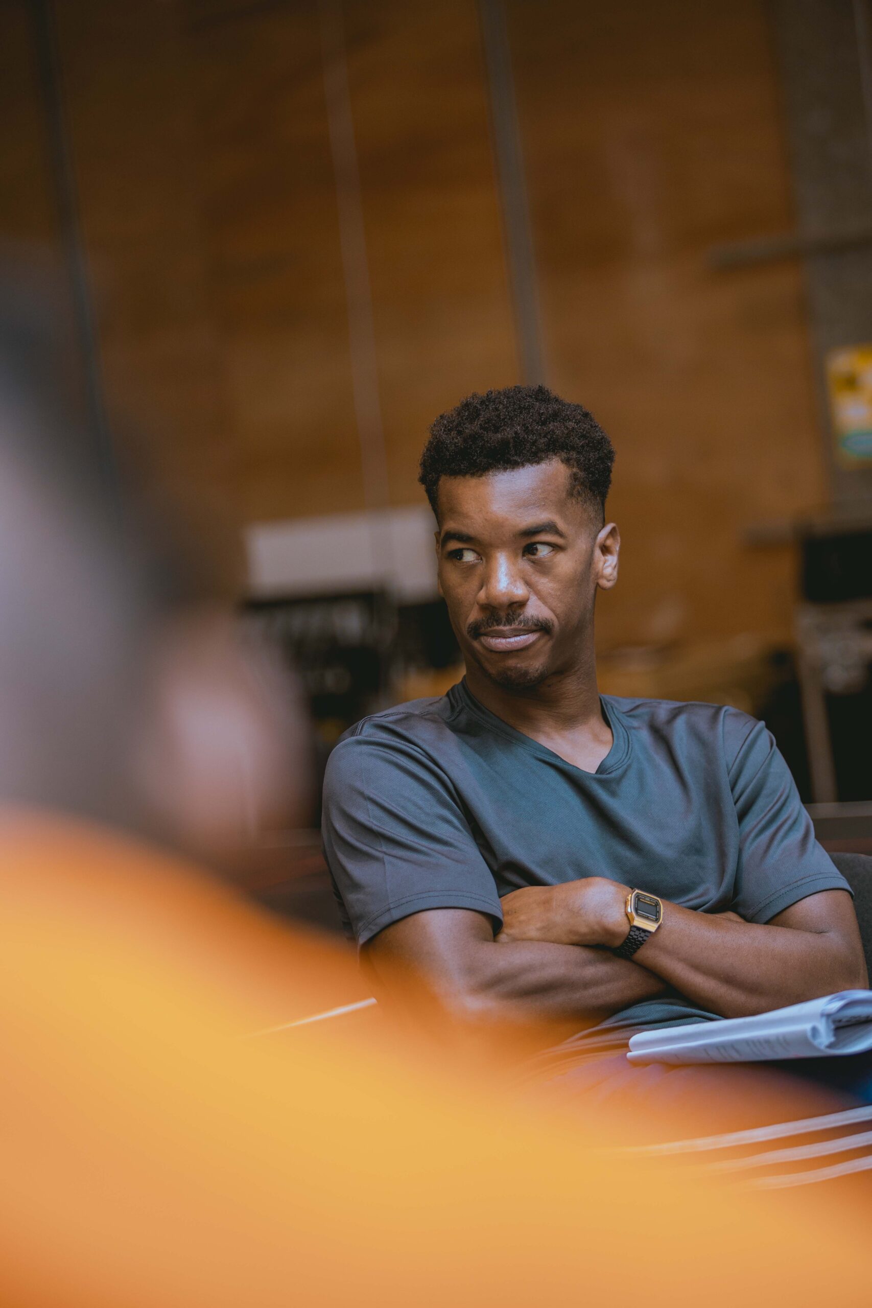 In a wood-cladded rehearsal room, Troy Glasgow (Father) is seated with his arms crossed. He is wearing a grey t-shirt, and is observing something to his right.